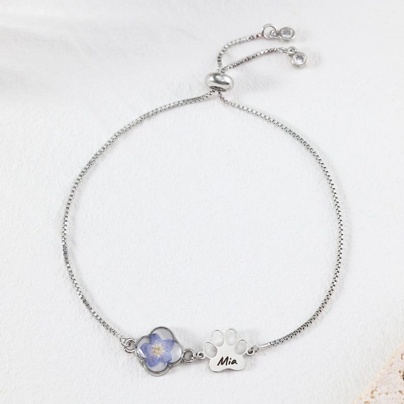 Puede incluir: Pulsera ajustable plateada. Presenta un tr&eacute;bol de cuatro hojas con una flor azul y un dije de huella de pata con el nombre "Mia". La pulsera tiene una cadena delicada y un cierre deslizante.