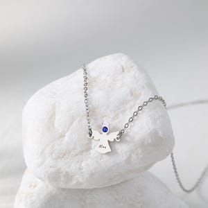 May include: A silver angel pendant necklace with a blue gemstone halo and a delicate chain. The angel has outstretched wings and the name "Max" engraved on the front. The necklace is displayed on a white stone.