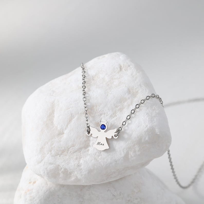 May include: A silver angel pendant necklace with a blue gemstone halo and a delicate chain. The angel has outstretched wings and the name "Max" engraved on the front. The necklace is displayed on a white stone.