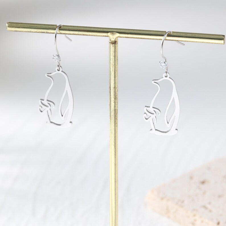 May include: A pair of silver-colored penguin earrings. Each earring features a cutout design of a penguin with a small, dangling accent at the top. The earrings are displayed on a gold-colored jewelry stand against a soft, neutral background.