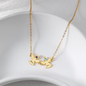 May include: A gold-colored necklace featuring two angel pendants, each with a gemstone and a name. The pendant on the left has a purple gemstone and the name "Milo", while the right pendant has a blue gemstone and the name "Max".