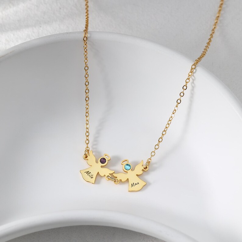 May include: A gold-colored necklace featuring two angel pendants, each with a gemstone and a name. The pendant on the left has a purple gemstone and the name "Milo", while the right pendant has a blue gemstone and the name "Max".