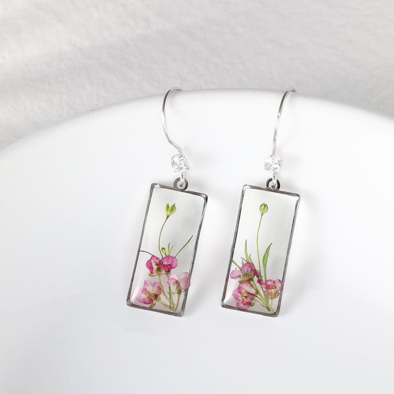 May include: A pair of rectangular earrings with a clear resin center, showcasing pink and green dried flowers. The earrings have silver-colored metal frames and a small, clear gem at the top of each hook.