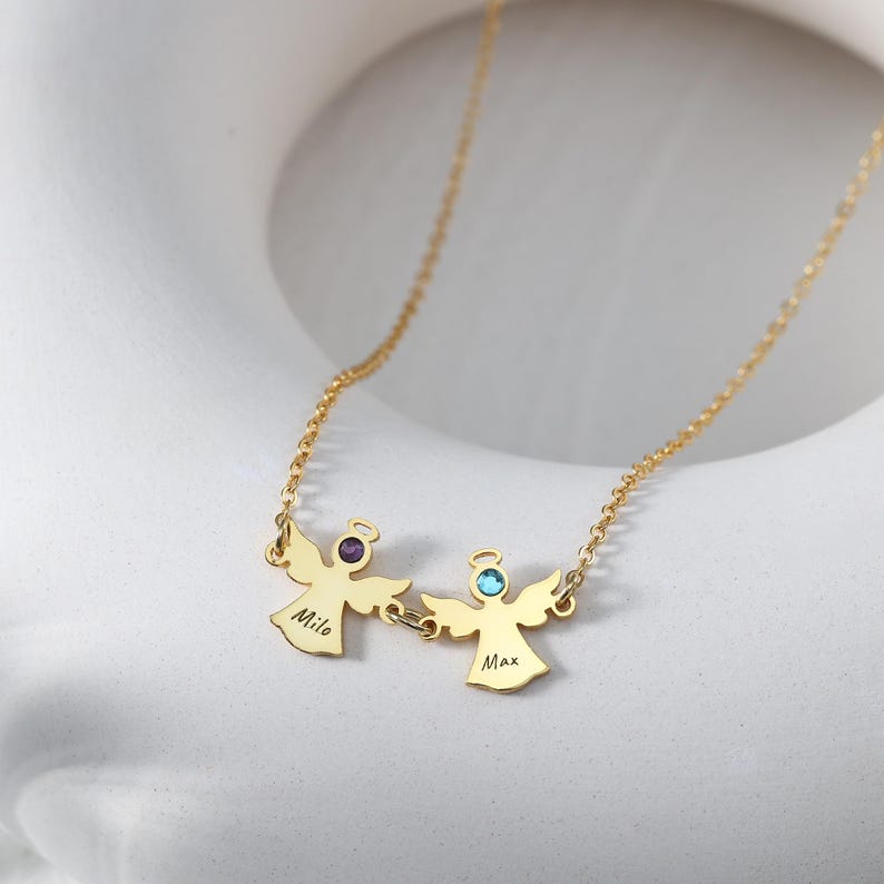 May include: A gold-colored necklace featuring two angel pendants. Each angel has a gemstone and a name engraved on it. One angel has a purple gemstone and the name "Milo", while the other has a blue gemstone and the name "Max".