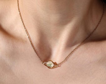 May include: A delicate gold-toned necklace featuring a small, oval pendant with a preserved white flower. The chain is fine and the pendant is set in a gold-colored bezel. The necklace is worn around the neck.