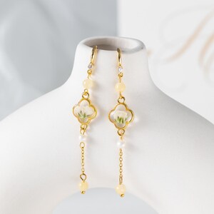 May include: Gold-toned dangle earrings with a four-leaf clover design. Each earring features a small white flower encased in clear resin, accented with yellow beads, a small diamond, and a dangling chain with a pearl and a yellow bead.