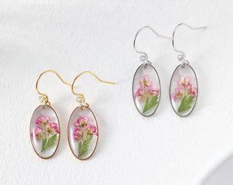 Könnte beinhalten: Ovale Ohrringe mit echten rosa Blüten und grünen Blättern, die in klarem Harz eingeschlossen sind. Ein Paar hat goldfarbene Haken, das andere silberfarbene. Die Ohrringe werden auf einer weißen Oberfläche präsentiert.