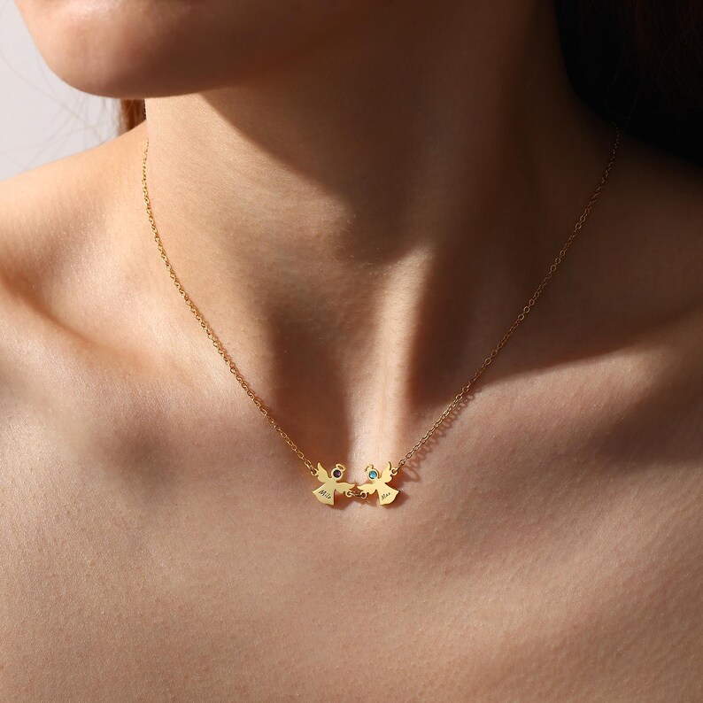 May include: A gold-colored necklace featuring two angel pendants. Each angel has a small gemstone and holds a heart. The necklace is worn around a person's neck.