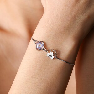 Puede incluir: Una pulsera plateada con un dije en forma de flor con un dise&ntilde;o floral morado claro y un dije de huella de pata con el nombre "Mia" grabado. La pulsera se lleva en la mu&ntilde;eca.