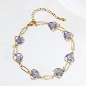 May include: A gold-toned bracelet featuring heart-shaped charms, each encasing a delicate blue flower. The bracelet's design incorporates oval links connecting the heart charms, creating a unique and elegant accessory. The clasp is adjustable.