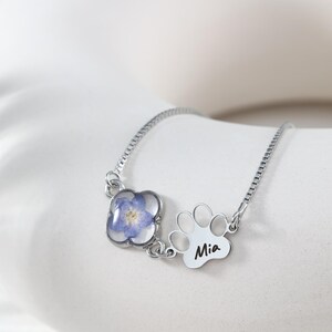 Puede incluir: Una pulsera de plata con un colgante de flor azul y un charm de huella de pata con el nombre "Mia" grabado. La flor est&aacute; encapsulada en resina transparente. La pulsera se muestra sobre una superficie blanca.