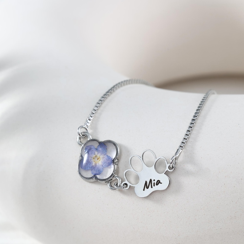 Puede incluir: Una pulsera de plata con un colgante de flor azul y un charm de huella de pata con el nombre "Mia" grabado. La flor est&aacute; encapsulada en resina transparente. La pulsera se muestra sobre una superficie blanca.