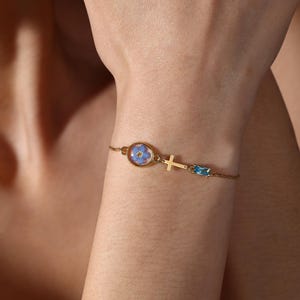 May include: A gold-colored bracelet featuring a small, oval-shaped pendant with a blue flower encased within. The bracelet also includes a small cross charm and a rectangular blue gemstone. The bracelet is delicate and elegant.
