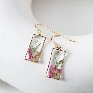 May include: A pair of gold-colored earrings with rectangular clear resin pendants. Each pendant contains pink and green dried flowers. The earrings have a hook design with a small clear gem near the ear. The earrings are displayed on a white surface.