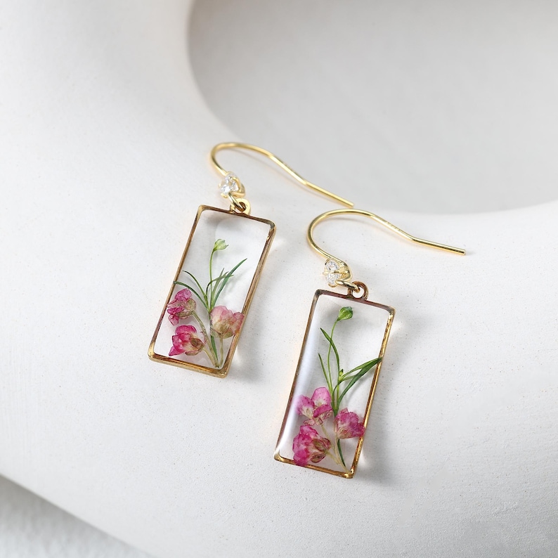 May include: A pair of gold-colored earrings with rectangular clear resin pendants. Each pendant contains pink and green dried flowers. The earrings have a hook design with a small clear gem near the ear. The earrings are displayed on a white surface.