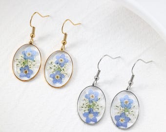 Puede incluir: Pendientes colgantes ovalados con flores azules y blancas prensadas, encapsuladas en resina transparente. Se muestran dos pares de pendientes, uno con ganchos y marcos dorados, el otro con ganchos y marcos plateados. Sobre una superficie blanca.