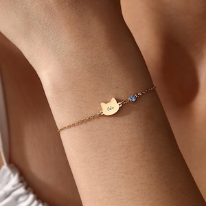 Puede incluir: Pulsera dorada con un dije en forma de gato con el nombre "Odin" grabado. Una pequeña gema azul está engastada cerca del dije. La pulsera es delicada y se lleva en la muñeca.