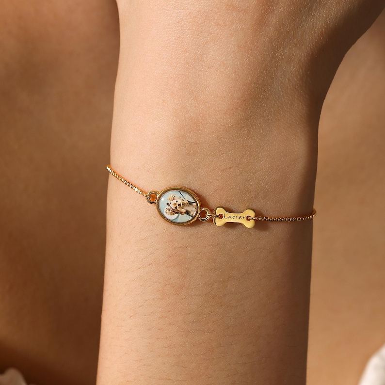 Op de afbeelding: Goudkleurige armband met een ovale bedel met een hondenportret en een botvormige bedel met de naam "Caesar". De armband heeft een fijne ketting.