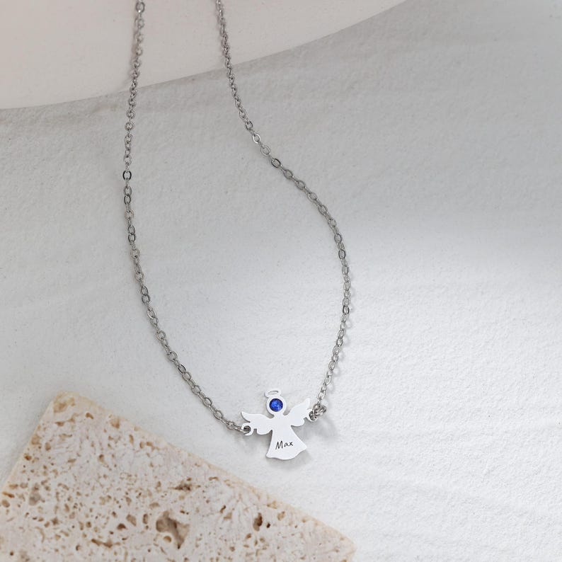May include: A silver-colored necklace featuring a pendant shaped like an angel with a blue gemstone. The angel pendant has the name "Max" engraved on it. The necklace is displayed on a light-colored surface.