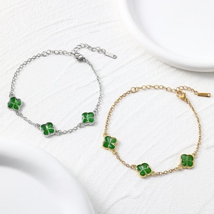 May include: Two bracelets with a clover design. One is silver-toned, the other gold-toned. Each bracelet has three green clover charms. The bracelets are shown on a white surface.