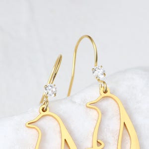 May include: Gold-colored dangle earrings featuring a penguin silhouette design. Each earring has a small, clear gemstone at the top and bottom of the penguin shape. The earrings are suspended from a curved hook.