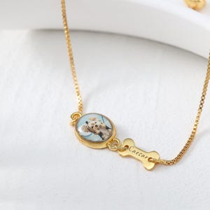 Op de afbeelding: Goudkleurige armband met hondenthema. De armband heeft een kleine, ovale hanger met een foto van een hond. Een botvormige bedel met de naam "Caesar" is aan de hanger bevestigd. De ketting is een fijne boxketting.