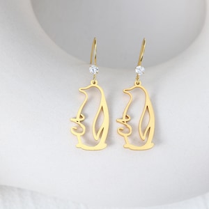 May include: A pair of gold-colored penguin-shaped earrings. Each earring features a dangling penguin outline with a small, clear gemstone at the top. The earrings have a simple, elegant design.