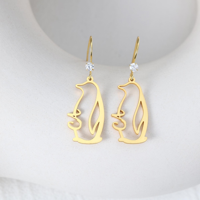 May include: A pair of gold-colored penguin-shaped earrings. Each earring features a dangling penguin outline with a small, clear gemstone at the top. The earrings have a simple, elegant design.