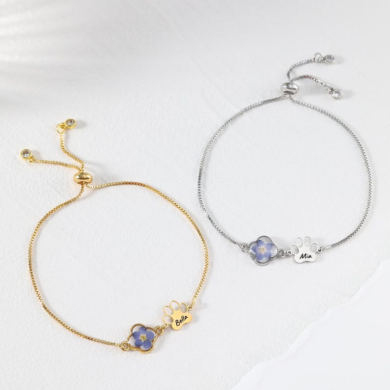 Puede incluir: Dos pulseras ajustables, una dorada y otra plateada. Cada una presenta una peque&ntilde;a flor azul y un dije de huella de pata. La pulsera dorada tiene grabado "Bella", y la plateada tiene "Mia".