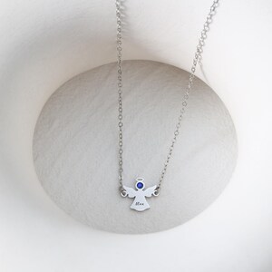 May include: A silver-colored necklace featuring an angel pendant. The pendant has a blue gemstone halo and the name "Max" engraved on it. The necklace is displayed on a white, textured surface.