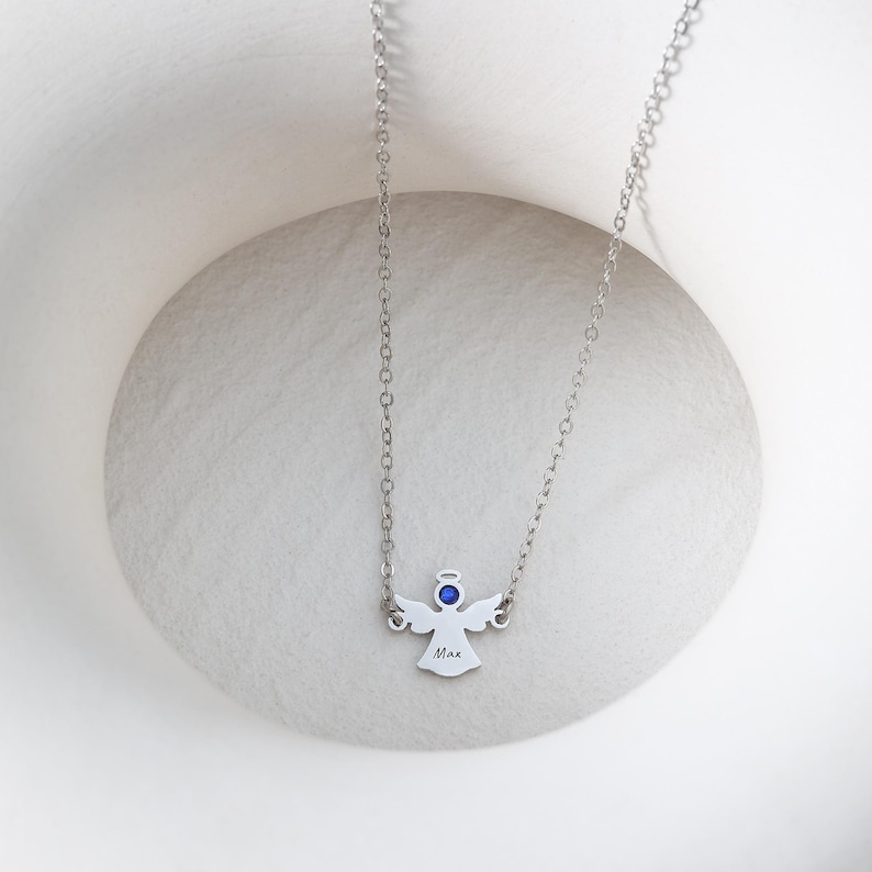 May include: A silver-colored necklace featuring an angel pendant. The pendant has a blue gemstone halo and the name "Max" engraved on it. The necklace is displayed on a white, textured surface.