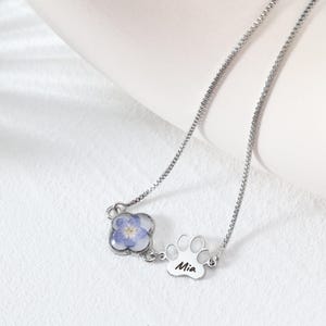 Puede incluir: Un collar plateado con un colgante de tr&eacute;bol de cuatro hojas con una flor azul y un dije de huella de pata. El dije de huella de pata tiene el nombre "Mia" grabado. El collar est&aacute; sobre una superficie blanca.