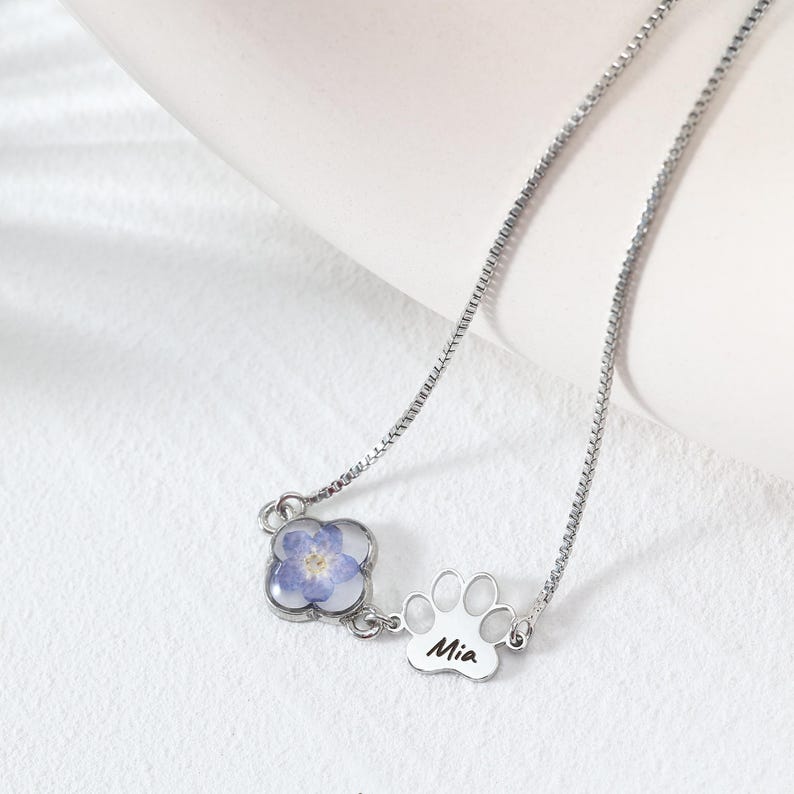 Puede incluir: Un collar plateado con un colgante de tr&eacute;bol de cuatro hojas con una flor azul y un dije de huella de pata. El dije de huella de pata tiene el nombre "Mia" grabado. El collar est&aacute; sobre una superficie blanca.