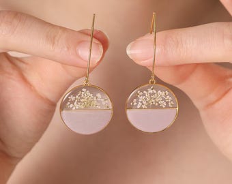 Puede incluir: Pendientes colgantes dorados con diseño circular. Los pendientes presentan una sección superior de resina transparente llena de pequeñas flores blancas y una sección inferior rosa claro. Los pendientes están sostenidos por un fino alambre dorado.