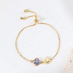 Puede incluir: Pulsera dorada ajustable con una flor azul y un dije de huella de pata con el nombre "Bella". La pulsera tiene una cadena delicada y una cuenta deslizante para ajustar. La pulsera est&aacute; sobre una superficie blanca.