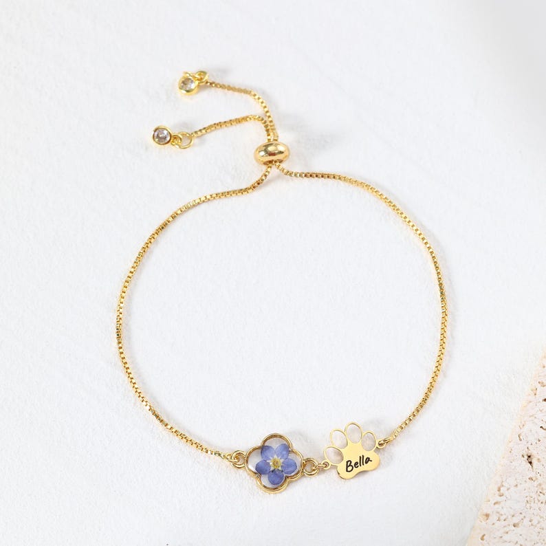 Puede incluir: Pulsera dorada ajustable con una flor azul y un dije de huella de pata con el nombre "Bella". La pulsera tiene una cadena delicada y una cuenta deslizante para ajustar. La pulsera est&aacute; sobre una superficie blanca.