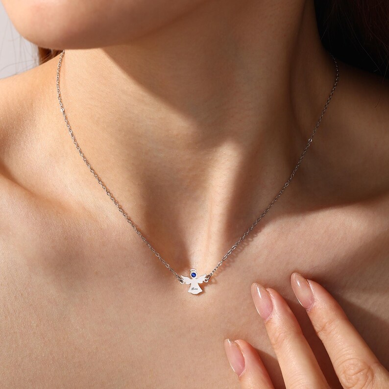 May include: A silver necklace featuring a small angel pendant with a blue gemstone eye. The angel has outstretched wings and a delicate chain. The necklace is worn around the neck.