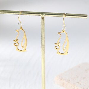 May include: A pair of gold-colored penguin earrings. Each earring features a cutout design of a penguin holding a small object. The earrings hang from a gold-colored hook with a small, clear gem.