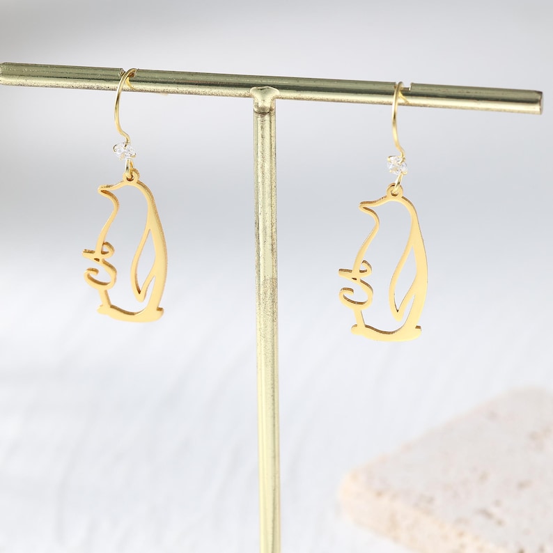 May include: A pair of gold-colored penguin earrings. Each earring features a cutout design of a penguin holding a small object. The earrings hang from a gold-colored hook with a small, clear gem.