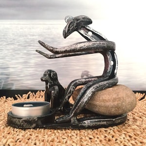 Puede incluir: Un portavelas rústico de metal que representa una figura sentada con un perro, una piedra y un portavelas. La escultura tiene un acabado plateado envejecido, creando una estética vintage. El portavelas está colocado sobre una estera tejida.