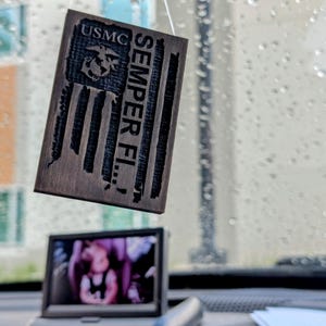 May include: A wooden USMC flag-shaped car air freshener with the words "USMC" and "Semper Fi" engraved. The air freshener hangs in front of a small screen displaying a photo. The background shows a window with raindrops.