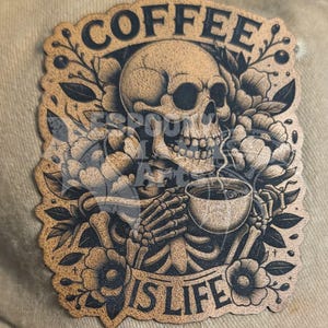 Puede incluir: Gorra de béisbol beige con un parche decorativo que muestra una calavera sosteniendo una taza de café. El parche tiene las palabras "COFFEE IS LIFE" y está rodeado de diseños florales y de hojas. La calavera y los elementos florales son en tonos marrones y negros.