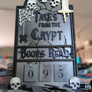 May include: A gray and black "Tales From The Crypt" book counter with a spider web, skulls, and a candle. The counter reads "095". The product is a decorative item.