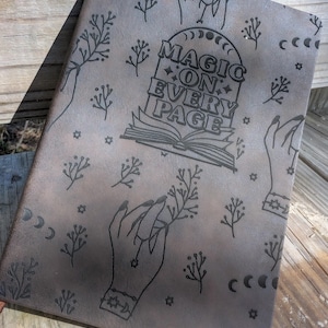 May include: A gray leather journal with a black embossed design. The design features hands, stars, moons, and the text "Magic on Every Page".