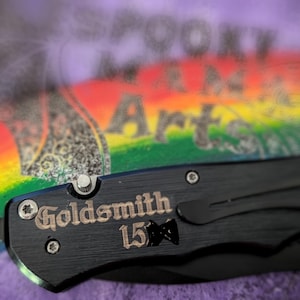 以下が含まれることがあります： 黒い折りたたみナイフ。ハンドルには「Goldsmith 15」の文字が刻印されています。ナイフには黒いポケットクリップとシルバーのボタンが付いています。背景には虹と「Spooky Mama Arts」の文字があります。