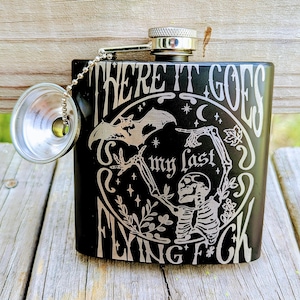 Könnte beinhalten: Schwarze Edelstahl-Taschenflasche mit silbernem Schraubverschluss und angebrachtem Becher. Die Flasche hat ein kreisförmiges Design mit einem Skelett, einer Fledermaus und dem Schriftzug "WHERE IT GOES my last FLYING F*CK".