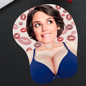 May include: Ergonomic mousepad featuring a woman's face and a blue bra design. The background is decorated with red lipstick kiss marks. The mousepad is on a black desk next to a laptop, red pencil, and a red notebook.