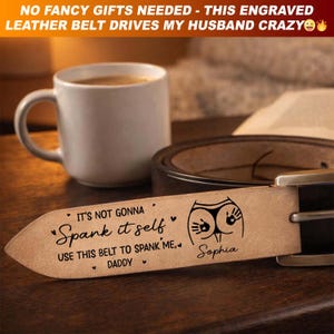 May include: A brown leather belt with a silver buckle. The belt is engraved with the text "It's not gonna Spank it self, Use this belt to spank me, Daddy" and a graphic. A white mug and a cup of coffee are in the background.