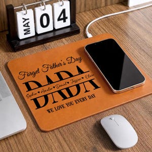 May include: A brown leather desk mat with the text "Forget Father's Day DADA" and the names Sophia, Amelia, Daniel, Jessica, and Olivia. A black smartphone, white mouse, and a wooden calendar are on the desk.
