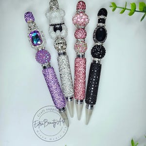 May include: Four decorative pens with jewel-encrusted barrels and ornate tops. The pens feature a variety of colors, including purple, silver, pink, and black. The tops include a variety of shapes and designs, including a bow tie, and a large oval gem. The pens are labeled "Extra Beauty Art."